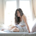 Young woman sitting on a bed holding her stomach in discomfort, appearing to experience pain or discomfort.