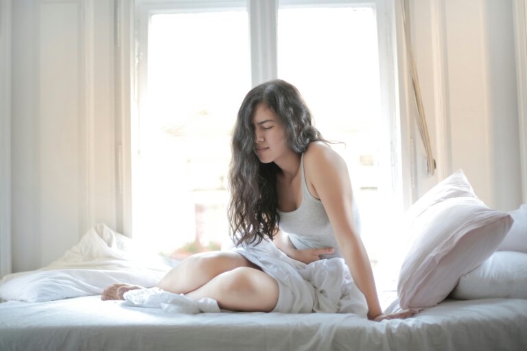 Young woman sitting on a bed holding her stomach in discomfort, appearing to experience pain or discomfort.