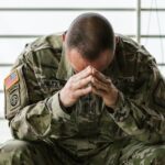 A soldier in a camouflage uniform sits with his head bowed and hands clasped in front of his face, appearing distressed.