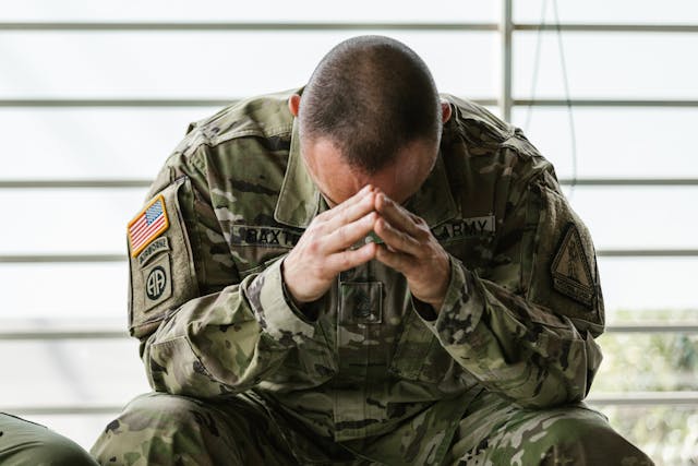 A soldier in a camouflage uniform sits with his head bowed and hands clasped in front of his face, appearing distressed.