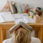 A person sitting at a desk with a book resting on their head, symbolizing mental overload, anxiety, and the weight of fear.