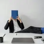 Leader covering face with notebook at desk representing imposter syndrome and workplace anxiety